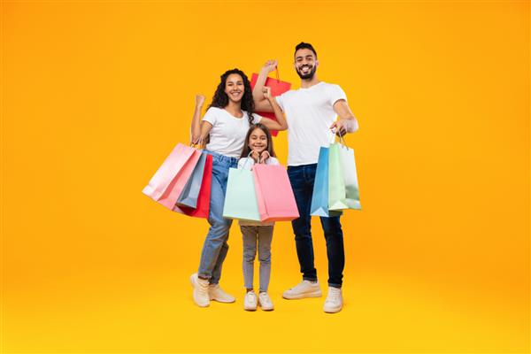 خانواده سه نفره عرب در حال خرید با هم در حالی که کیسه‌های کاغذی رنگارنگ را در دست دارند که روی پس‌زمینه زرد ایستاده‌اند استودیو عکس والدین آگهی خرید لباس جدید دختر طول کامل