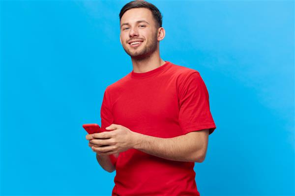 مرد خوش تیپ برنزه خندان و شاد با تی شرت قرمز پیام در حال خرید آنلاین با استفاده از ژست تلفنی جدا شده در پس زمینه آبی استودیو کپی فضای بنر موکاپ مفهوم سبک زندگی افراد آنلاین