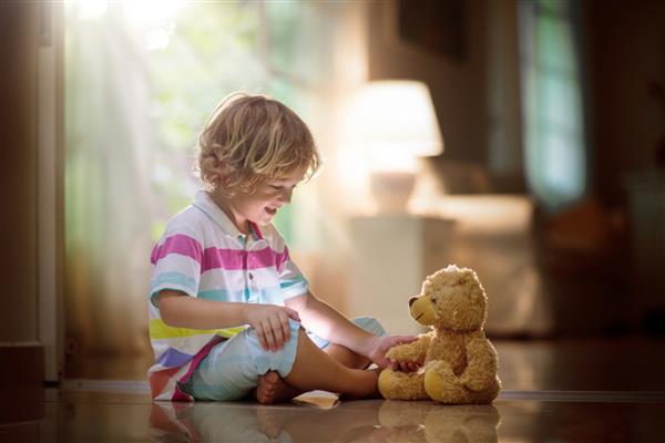 بازی کودک با خرس عروسکی پسر بچه ای که اسباب بازی مورد علاقه اش را در آغوش گرفته است بچه و حیوان عروسکی در خانه کودک نوپایی که هنگام غروب خورشید روی کف اتاق نشیمن با پنجره بزرگ نشسته است بچه ها در داخل خانه بازی می کنند
