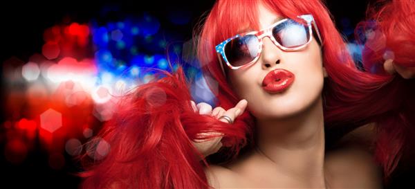 زن زیبای سکسی با موهای قرمز بلند با عینک آفتابی پرچم آمریکا چهارم ژوئیه را جشن می گیرد و لب های پرپشت خود را در یک حرکت حسی به دوربین می زند