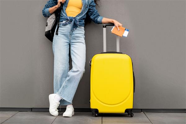 زن ناشناخته با لباس مسافرتی شیک و گاه به گاه که روی پس‌زمینه خاکستری ایستاده است چمدان و کوله‌پشتی زرد را حمل می‌کند پاسپورت و بلیط هواپیما را در دست دارد برش داده شده فضای کپی