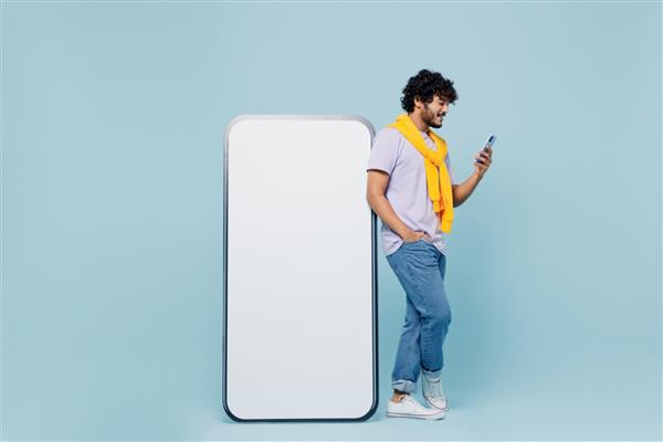 مرد جوان ریش دار هندی 20 ساله تی شرت سفید را در نزدیکی تلفن همراه بزرگ با صفحه نمایش خالی می پوشد که روی پرتره استودیویی با پس زمینه آبی روشن ساده جدا شده است
