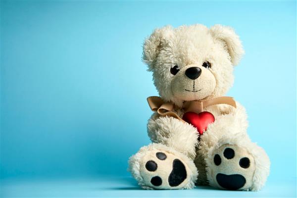 خرس عروسکی قلب را روی پس‌زمینه آبی با فضای کپی نگه می‌دارد اسباب بازی خرس کرکی سفید با قلب قرمز مفهوم روز عشق و ولنتاین