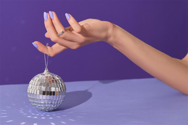 دست های زن آراسته که توپ دیسکو را در پس زمینه بنفش نگه می دارند مفهوم سالن زیبایی مانیکور پدیکور