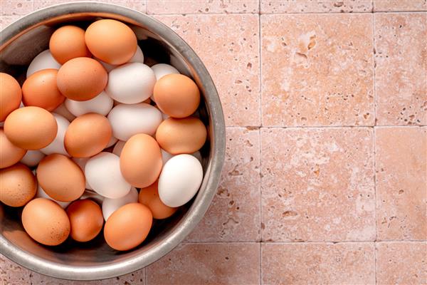 تخم مرغ در کاسه فلزی در پس زمینه بژ با فضای کپی نمای بالای تخم مرغ قهوه ای خام و تخم مرغ سفید با پر مرغ