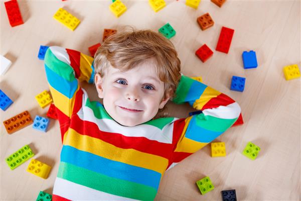 کودک بور کوچک در حال بازی با تعداد زیادی بلوک پلاستیکی رنگارنگ در داخل خانه پسر بچه ای که پیراهن رنگارنگ پوشیده و با ساختن و خلق کردن سرگرم می شود