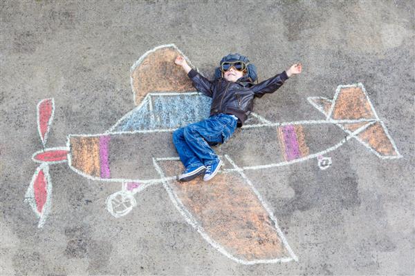 پسر بچه کوچک شاد با لباس خلبانی که با نقاشی هواپیما با گچ رنگارنگ سرگرم می شود اوقات فراغت خلاقانه برای کودکان در فضای باز در تابستان