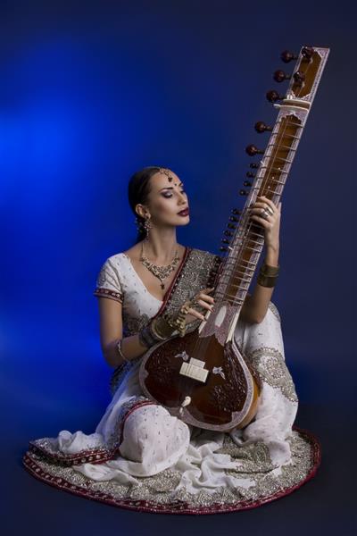 زن جوان هندی زیبا با لباس سنتی ساری با جواهرات شرقی آرایش عروس در حال نواختن سیتار دختر زیبای بالیوود افسانه شرقی اثر لنز آبی