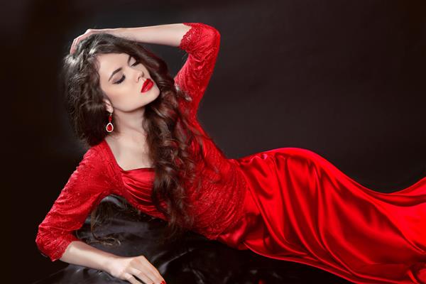 زن قرمزپوش در تاریکی دراز کشیده است مدل دخترانه وسوسه انگیز با لباس مجلسی سکسی با لب های حسی و موهای بلند جدا شده در زمینه مشکی