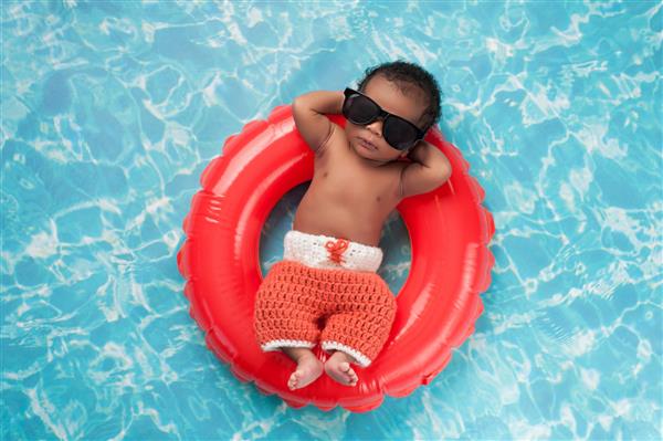نوزاد پسر دو هفته ای که روی یک حلقه شنا بادی کوچک می خوابد او شورت قلاب بافی و عینک آفتابی مشکی به چشم دارد