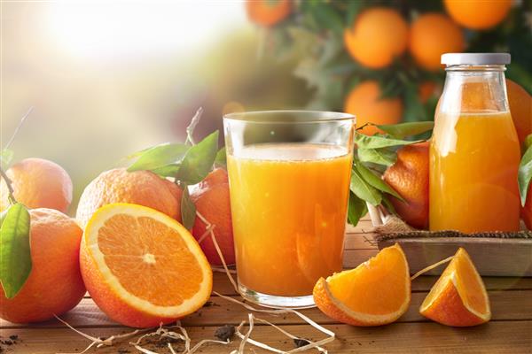 یک لیوان آب پرتقال روی یک میز چوبی با بطری و بخش های پرتقال پس زمینه درخت و مزرعه با خورشید عصر ترکیب افقی نمای جلویی