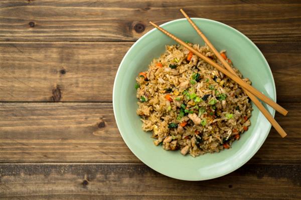 برنج سرخ شده با مرغ ریحان با چاپستیک در پس زمینه چوبی که از بالا مشاهده می شود این غذای الهام گرفته از تایلندی برای یک ناهار سریع یا به عنوان غذای جانبی مناسب است