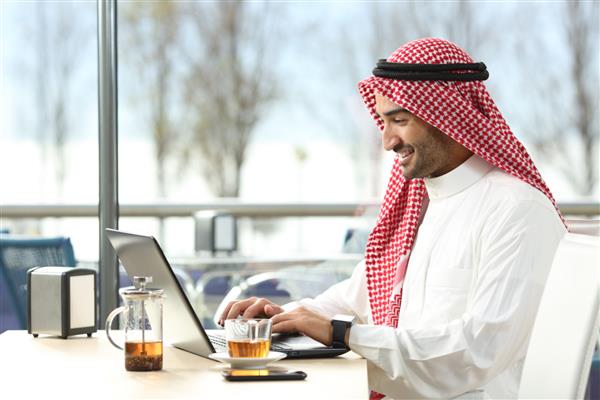 مرد عرب سعودی در حال کار آنلاین با لپ‌تاپ و ساعت هوشمند در کافی‌شاپ یا بار هتل با پنجره و تراس در فضای باز در پس‌زمینه