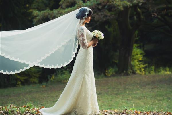 نمای کناری تمام قد از یک عروس جوان سبزه شهوانی با لباس عروسی سفید بلند و مقنعه ایستاده در جنگل و دسته گل در فضای باز در پس زمینه طبیعی تصویر افقی