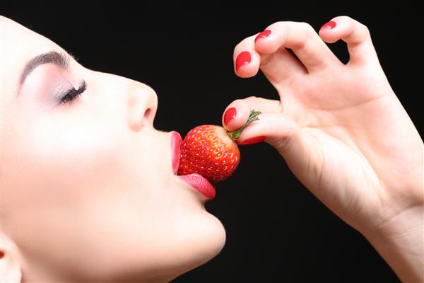 زن جوان شهوانی در حال خوردن توت فرنگی در پس زمینه سیاه نزدیک