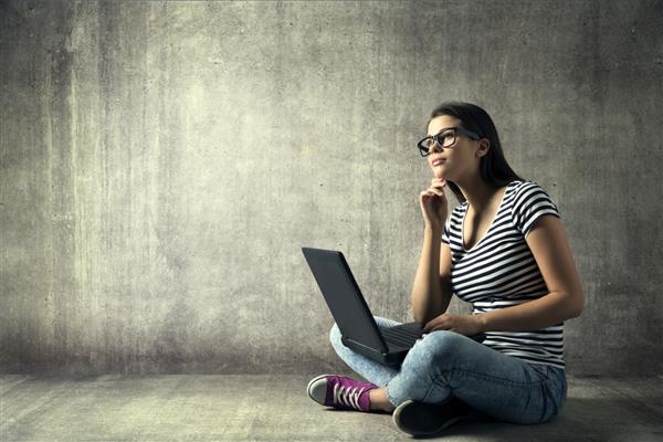 زن در حال استفاده از لپ تاپ دختر جوان با عینک در حال فکر کردن روی نوت بوک دانش آموز با کامپیوتر که روی پس زمینه طبقه گرانج نشسته است