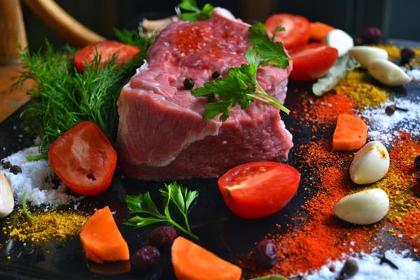 استیک گوشت گاو خام تازه روی زمینه سیاه با هویج پراکنده گوجه فرنگی سیر سبزیجات نمک و ادویه جات ترشی جات