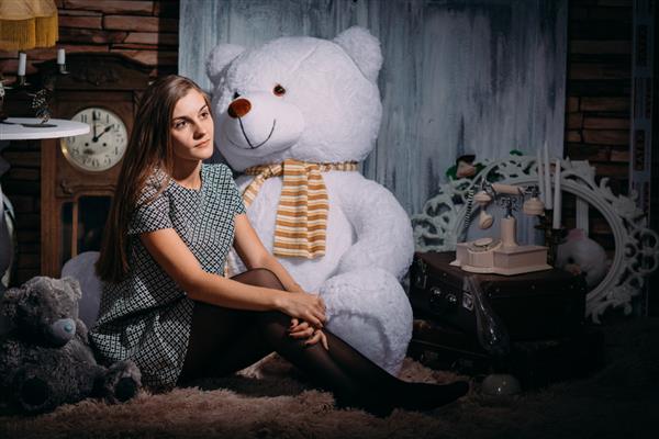 یک دختر سکسی زیبا با لباس کوتاه و جوراب شلواری روی زمین با یک خرس عروسکی بزرگ در یک استودیو عکس نشسته است