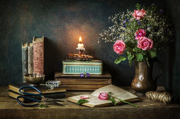 طبیعت بی جان کلاسیک با کتاب های قدیمی با گل های رز صورتی زیبا جعبه های قدیمی قیچی شمع نورانی نخ کرفس روی زمینه چوبی روستایی قرار داده شده است