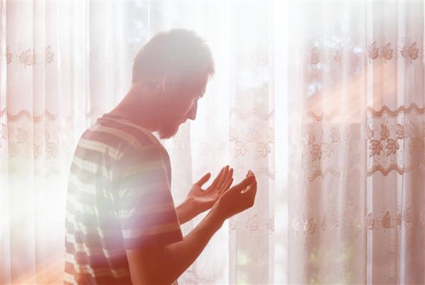 مرد جوان مسلمان با دستان بالا در حالت دعای هوا وقت نماز می گذراند پرتوهای نوری که از پنجره روشن بهشت می‌آیند