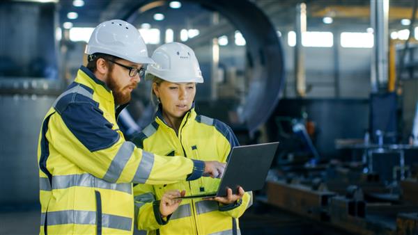 مهندسان صنعتی مرد و زن در کلاه های سخت در مورد پروژه جدید در حین استفاده از لپ تاپ بحث می کنند آنها ژست های نمایشی می دهند آنها در یک کارخانه تولیدی صنایع سنگین کار می کنند