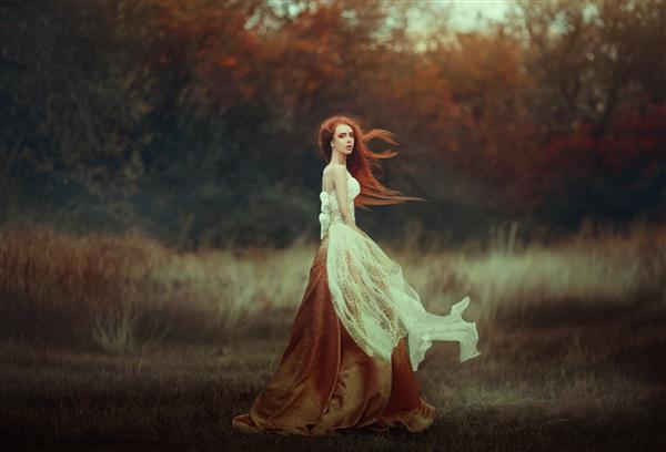 زن جوان زیبا با موهای قرمز بسیار بلند در لباس قرون وسطایی طلایی که در جنگل پاییزی قدم می زند موهای قرمز بلند در باد ایجاد می شوند