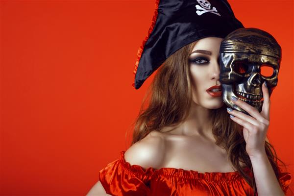 پرتره زن جذاب سکسی با آرایش تحریک آمیز در لباس دزدان دریایی که نیمی از صورت خود را پشت ماسک جمجمه پنهان کرده است جدا شده در پس زمینه قرمز روشن نزدیک فضای کپی