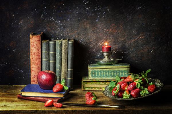 طبیعت بی جان کلاسیک با کتاب های قدیمی با بشقاب نقره ای پر از توت فرنگی تازه جعبه های قدیمی شمع نورانی و سیب قرمز در زمینه چوبی روستایی