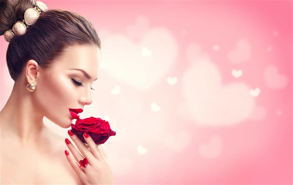 زن زیبایی روز ولنتاین با گل رز قرمز مدل چهره دخترانه پرتره با رز قرمز در دست لب و ناخن قرمز پس زمینه صورتی تار آرایش و مانیکور لاکچری زیبا مو