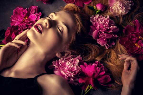پرتره زیبایی زن بلوند زیبا با لب‌های قهوه‌ای شهوانی که در میان گل‌های گل صد تومانی خوابیده است لوازم آرایشی آرایش عطرسازی