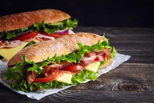 دو ساندویچ زیردریایی تازه با ژامبون پنیر بیکن گوجه فرنگی کاهو خیار و پیاز در زمینه چوبی تیره