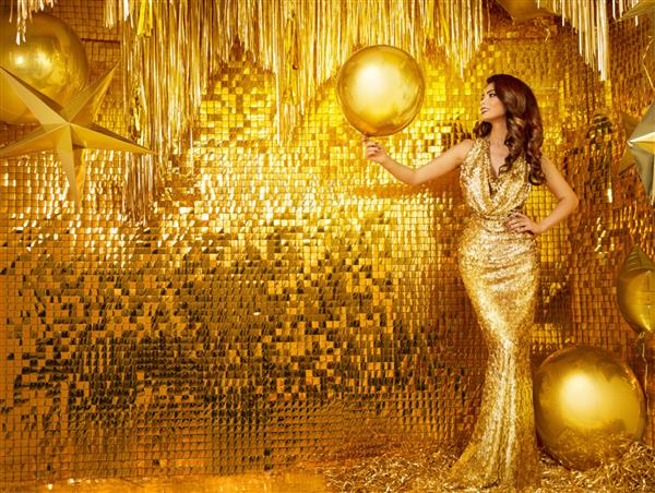 زنی با لباس مجلسی طلایی مدل مد لباس مجلسی بلند شب روی دیوار داخلی طلایی براق دختری شیک که بادکنک فویلی زرد در دست دارد جشن تولد را جشن می گیرد