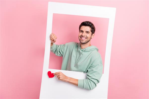 پرتره دست‌های فرد مثبت کارت پنجره کاغذی قلب کوچک را مانند جدا شده روی پس‌زمینه صورتی رنگ نگه می‌دارد
