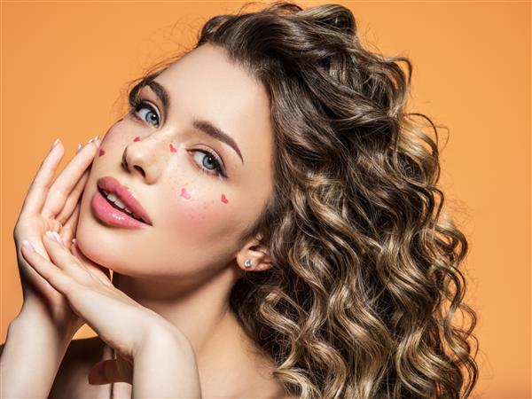 پرتره دختری زیبا با قلب های قرمز رنگ شده روی صورتش عکاسی مد بلوند با موهای مجعد یک مدل رسا و با آرایش روشن در استودیو ژست می گیرد