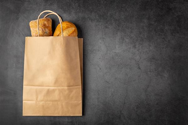 کیسه کاغذی یکبار مصرف از یک سوپرمارکت قابل بازیافت با نان های کامل پخته شده روی پس زمینه تیره با فضای کپی