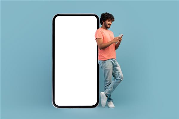 پسر هندی مجعد باحال با لباس گاه به گاه تکیه به تلفن هوشمند بزرگ با صفحه سفید سفید با استفاده از تلفن همراه و لبخند پس‌زمینه آبی استودیو ماکت پانوراما با فضای کپی