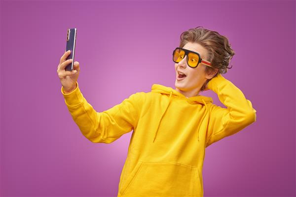 پسر نوجوان عاطفی بامزه ای با هودی زرد روشن که از طریق تماس ویدیویی با استفاده از گوشی هوشمند خود با خوشحالی صحبت می کند پس زمینه بنفش روشن نوجوانان سبک زندگی و ارتباطات نوجوانان مدرن