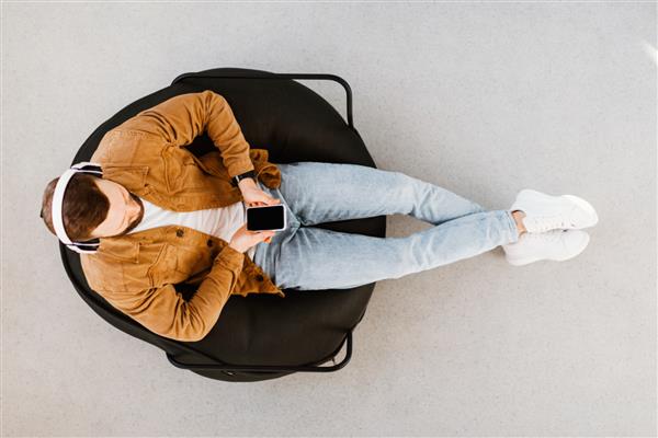 مردی با استفاده از پیامک و مرور اینترنت با گوشی هوشمند با هدفون گوش دادن به موسیقی آنلاین یا پادکست در داخل صندلی روی صندلی نشسته است عکس نمای بالا گجت ها و مفهوم فناوری