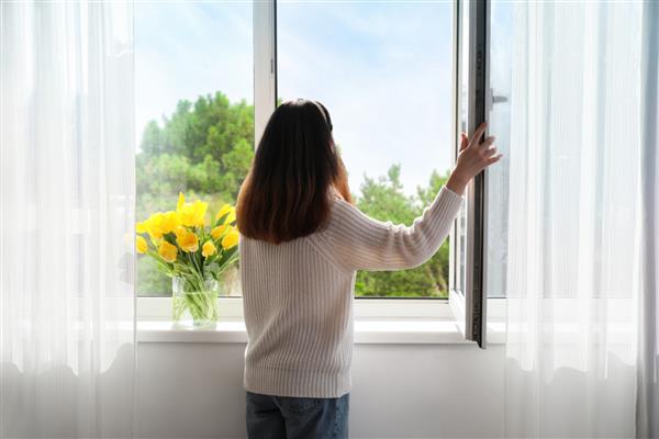 زن زیبای جوان آسیایی که پنجره خانه را در روز آفتابی باز می کند نمای پشت