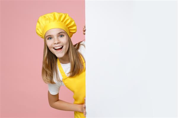 سرآشپز دختر نوجوان سرگرم‌کننده شیرینی‌پز خانه‌دار نانوا پیش بند زرد می‌پوشد کلاه تی‌شرت سفید تابلوی خالی را برای محتوای تبلیغاتی نگه دارید مکانی برای متن جدا شده در پس‌زمینه صورتی پاستلی مفهوم غذا