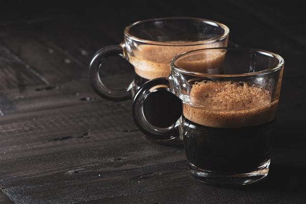 2 فنجان شیشه ای با اسپرسو روی سطح چوبی خاکستری و زمینه تیره قهوه تیره با کف قهوه ای در بالا