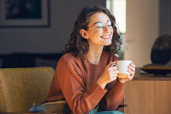 زن جوان شاد در حال نوشیدن یک فنجان چای در یک صبح پاییزی دختری رویایی که در اتاق نشیمن با فنجان قهوه داغ نشسته و زیر پتو با چشمان بسته لذت می برد زن زیبایی که در خانه ژاکت پوشیده است