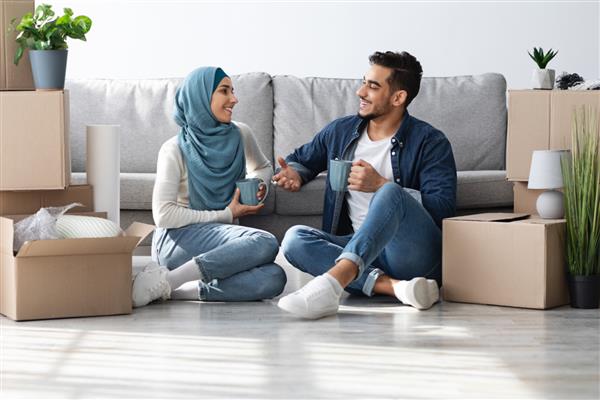 زن و شوهر جوان مثبت خاور میانه ای با حجاب نشستن روی زمین در اتاق خالی نوشیدن قهوه در حین باز کردن بسته بندی گفتگو و لبخند زدن برنامه ریزی برای بازسازی خانه