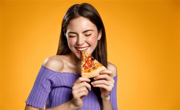 زن جوان شادی که در حال خوردن پیتزا ژست گرفتن با تکه‌های عشوه‌گری و خندیدن گاز گرفتن غذا مفهوم رستوران‌های فست فود پیتزا فروشی ایستادن روی پس‌زمینه نارنجی