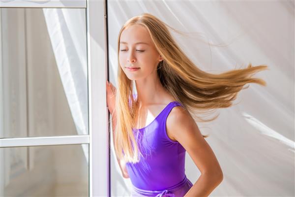 دختر بالرین جوان لاغر اندام با شلوار یاسی رنگی نزدیک پنجره باز نسیم پرده و موهای یک نوجوان را تکان می دهد