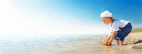 بازی بچه در ساحل در یک روز آفتابی گرم دختر کوچکی که لباس ملوانی پوشیده است با پای برهنه در ساحل شنی ایستاده و قایق را به دریا می اندازد کودک آرزوی سفر و ماجراجویی را دارد