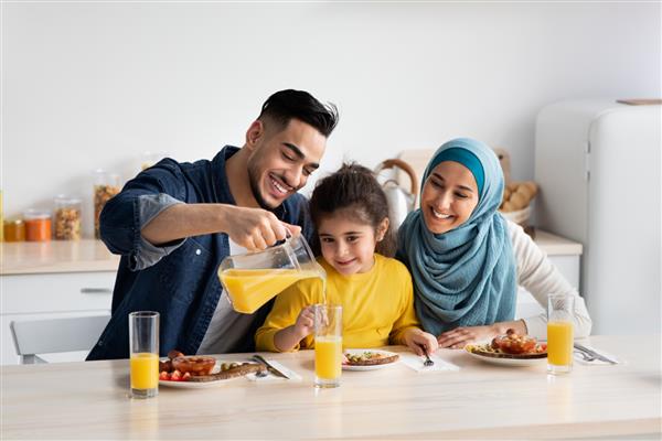 پرتره خانواده سه نفره خوشبخت خاورمیانه ای که با هم در آشپزخانه صبحانه می خورند والدین عرب با دختر کوچک ناز در حال لذت بردن از غذای خوشمزه و نوشیدن آب پرتقال در حالی که پشت میز نشسته اند