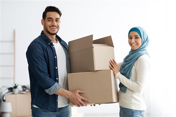 زن زیبا با حجاب به شوهرش جعبه های کاغذی با وسایلشان می دهد و به خانه جدید نقل مکان می کند خانواده شاد و جوان عرب دوست داشتنی که روز جابجایی دارند وسایل را بسته بندی می کنند یا در جعبه ها باز می کنند