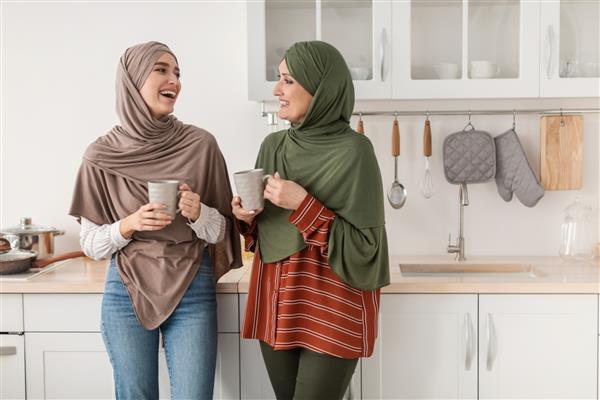 مادر مسلمان سالخورده شاد و دختر بالغ با حجاب و لذت بردن از قهوه صبحگاهی صحبت کردن و خندیدن با هم ایستاده در آشپزخانه مدرن در خانه مفهوم خانواده شاد خاورمیانه