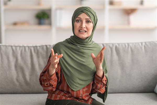 زن مسلمان میانسال با حجاب در حال صحبت با دوربین و نشسته روی مبل در خانه پرتره معلم بانوی خاورمیانه که از راه دور ارتباط برقرار می کند ارتباطات از راه دور مدرن تماس تصویری